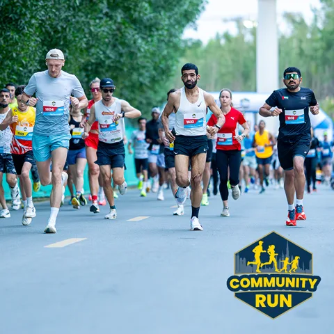 Runners participating in a community road race, wearing numbered bibs, with trees and a bridge in the background.