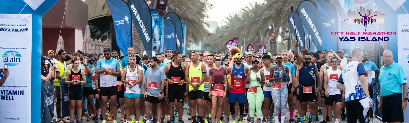 A diverse group of runners stands at the starting line of the City Half Marathon on Yas Island, surrounded by event banners and palm trees.