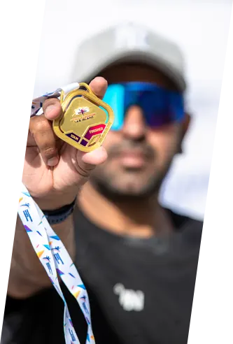 A person proudly holds up a shiny gold medal with a colorful ribbon, standing against a blurred background.