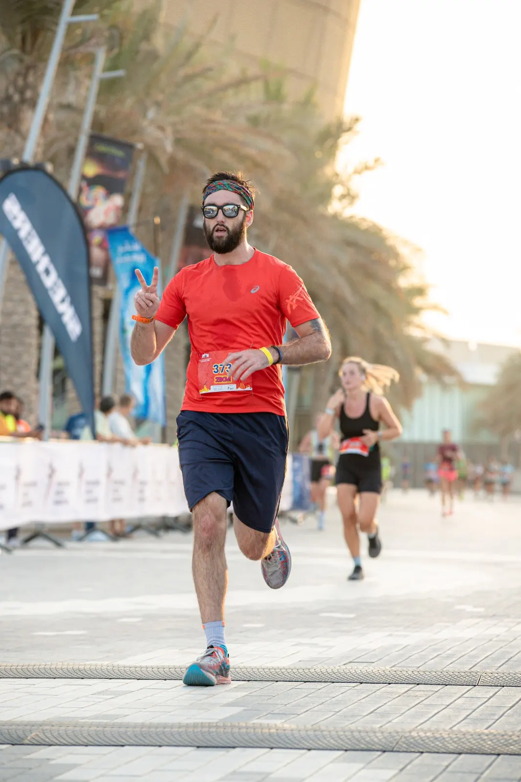 A runner in a red shirt and navy shorts participates in a race, flashing a peace sign while others run in the background.