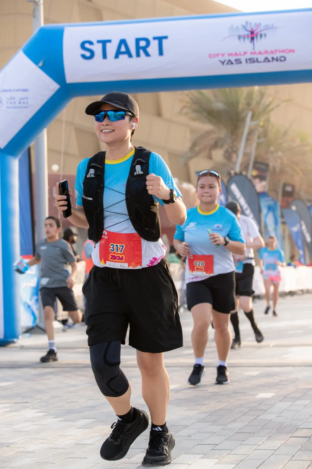 Runners participating in the City Half Marathon at Yas Island, passing under a blue START banner on a sunny day.
