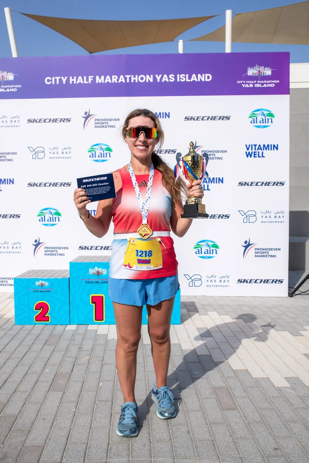 A runner proudly displays a trophy and a gift voucher, wearing a medal and sportswear, in front of a marathon backdrop with podiums.