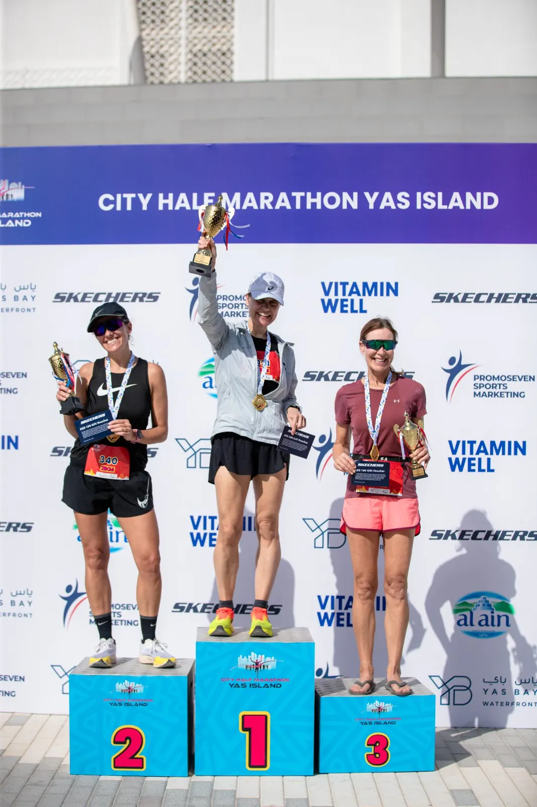 Three female marathon winners stand on podiums, holding trophies and certificates, celebrating their achievements at the Yas Island Half Marathon.