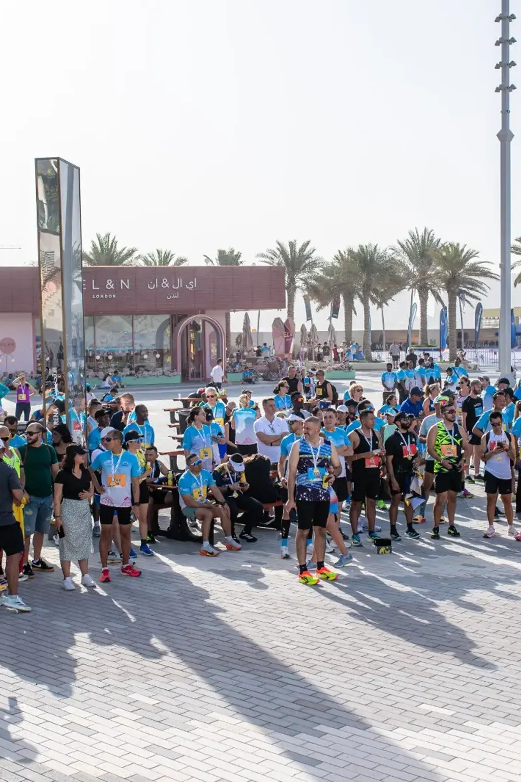 A large crowd of participants in blue shirts gathers outdoors at a lively event, with palm trees and tents in the background.