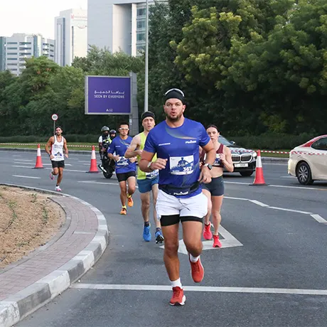 A group of runners in athletic gear races along a curved road with traffic cones and a backdrop of greenery and tall buildings.