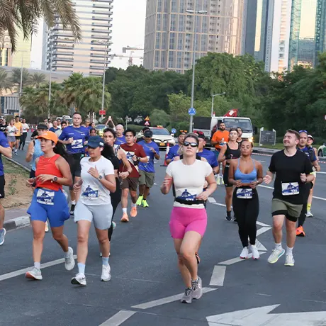 A lively group of runners in colorful athletic wear participates in an outdoor race, surrounded by urban scenery and greenery.