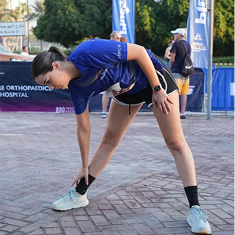 A person in a blue athletic shirt and black shorts stretches before a running event, surrounded by spectators and banners.