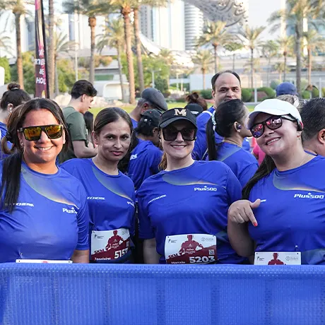 A group of participants in blue shirts, standing together with race bibs, at a sunny outdoor event surrounded by palm trees.