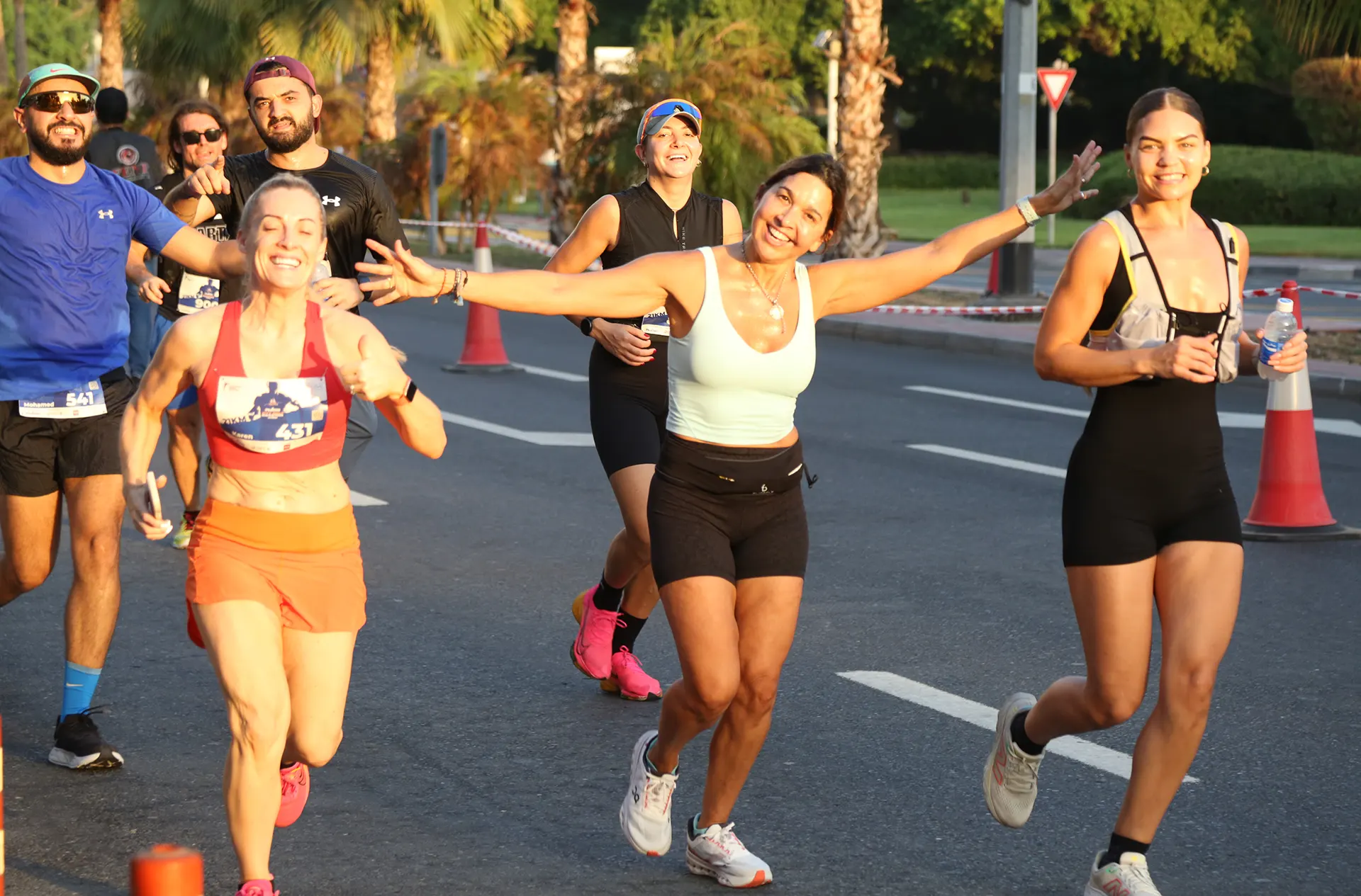 A group of runners participating in a race, with one woman joyfully extending her arms, wearing a light blue top and shorts