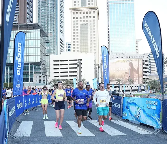 Runners in athletic gear cross a pedestrian lane, surrounded by tall buildings and banners, during an urban race event