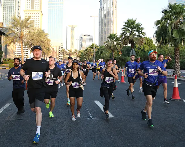 A group of runners in athletic gear participates in a race along a palm-lined urban street, with tall buildings in the background