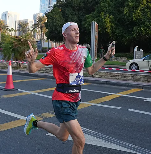 A runner in a colorful shirt holds a water bottle while sprinting on a city street, surrounded by palm trees and modern buildings
