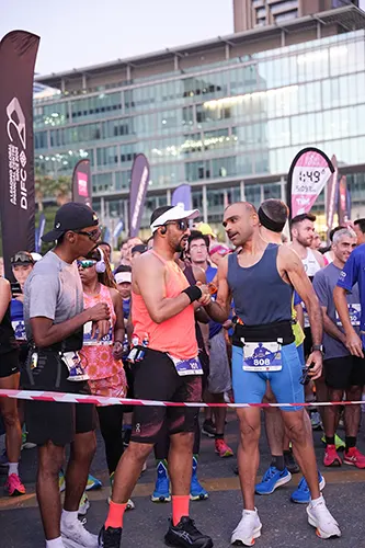 A group of male runners, dressed in colorful athletic wear, gather at the starting line of a race, with banners and a modern building in the background