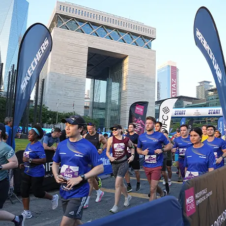 A group of runners in blue shirts participating in a race, passing a large building and banners at the starting line