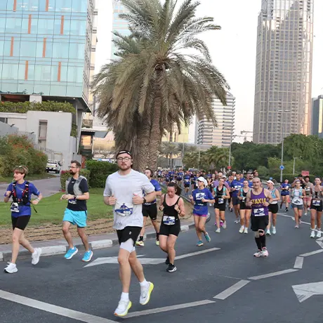 A large group of runners participates in a race along a city street, flanked by palm trees and modern skyscrapers