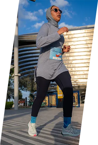 A runner in a gray outfit and black leggings strides confidently on a sunlit path, showcasing athletic gear and a race bib.