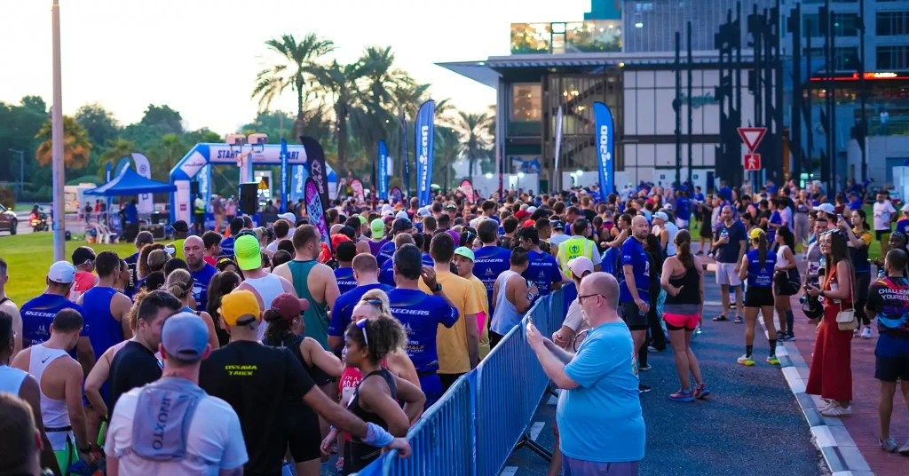 A large crowd of runners gathers at the starting line for an event, with banners and palm trees in the background.