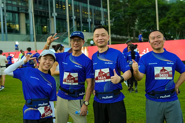 Four runners in blue shirts pose with smiles and hand gestures before a race, wearing bib numbers and fanny packs in a grassy area.