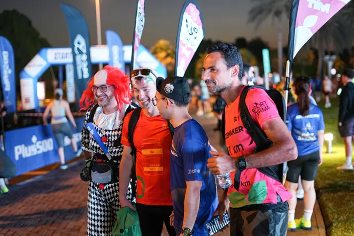 Four participants in colorful attire pose for a photo at a nighttime event, with banners and a festive atmosphere in the background.