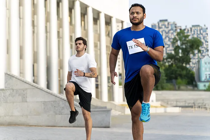 Two male runners perform high knees during an outdoor workout session near modern architecture, showcasing their athleticism and focus.