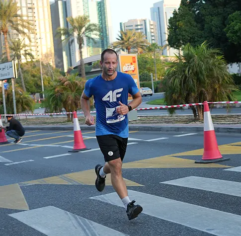 A runner in a blue shirt sprints across a pedestrian crossing, surrounded by palm trees and city skyscrapers in the background.