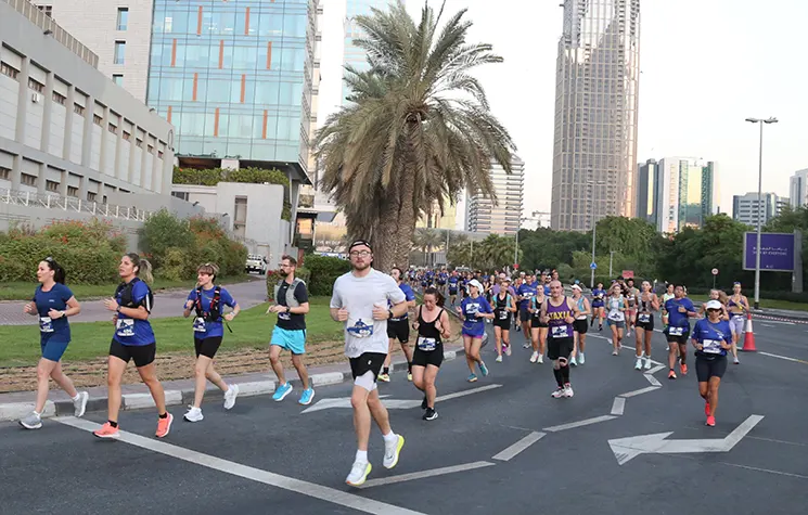 A group of diverse runners participates in a race, with palm trees and modern buildings in the background, showcasing a vibrant urban setting.