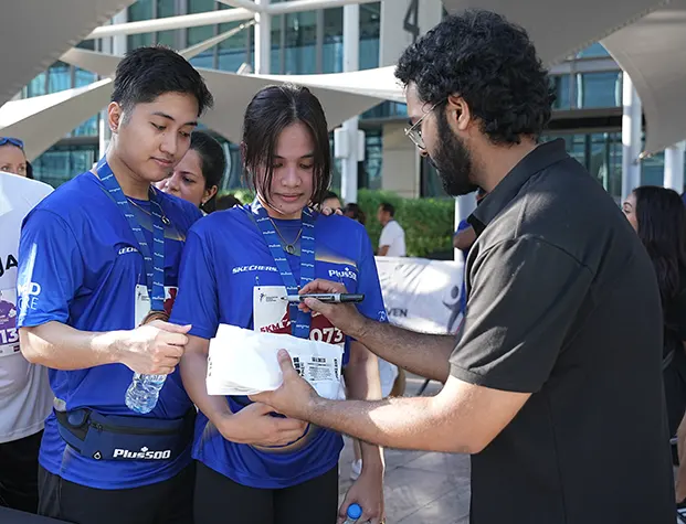 Participants in blue shirts gather for autographs after a race, holding water bottles and memorabilia in a lively outdoor setting.