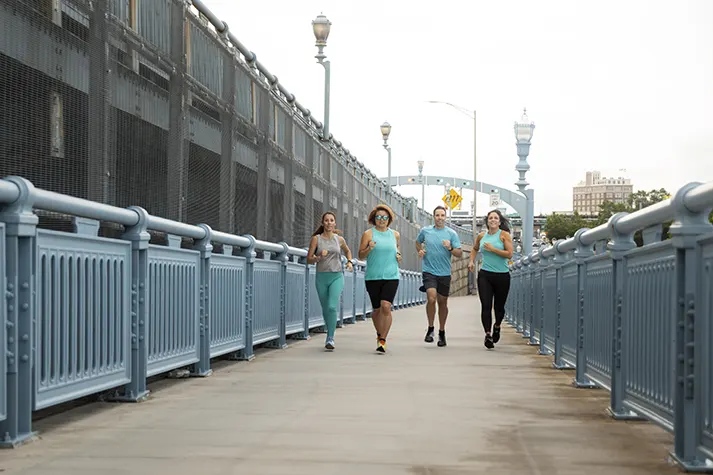 A diverse group of four individuals jogs along a blue railing pathway, set against an urban backdrop under a cloudy sky.