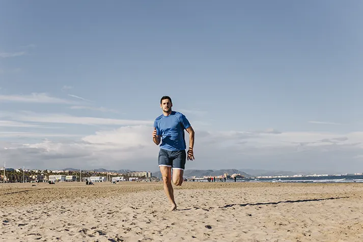 A person runs along a sandy beach with a clear blue sky and gentle waves in the background, showcasing an active, outdoor lifestyle.