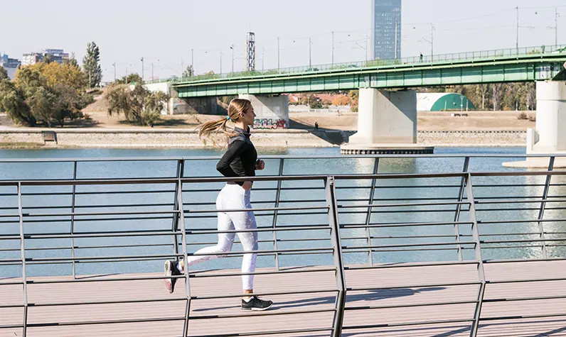 A woman jogs along a wooden pathway beside a river, with a green bridge in the background on a sunny day.
