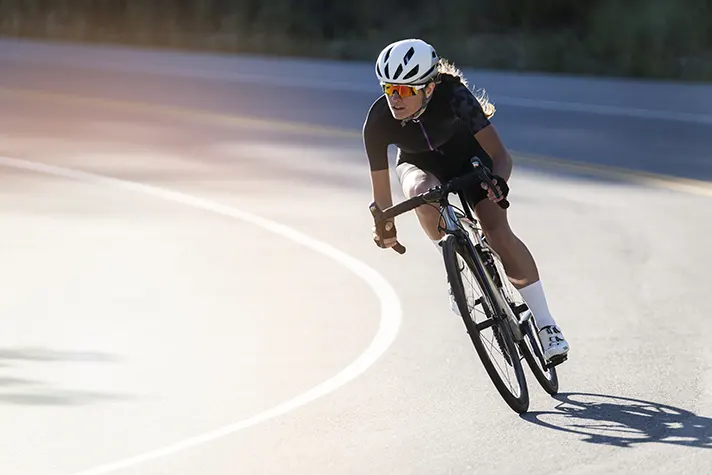 A cyclist leans into a sharp curve on a winding road, wearing a helmet and athletic gear, focused and in motion.