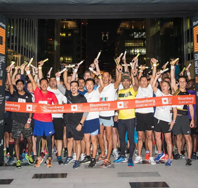 A group of runners celebrates at the finish line, holding relay batons and a ribbon that reads Bloomberg Square Mile Relay at night.