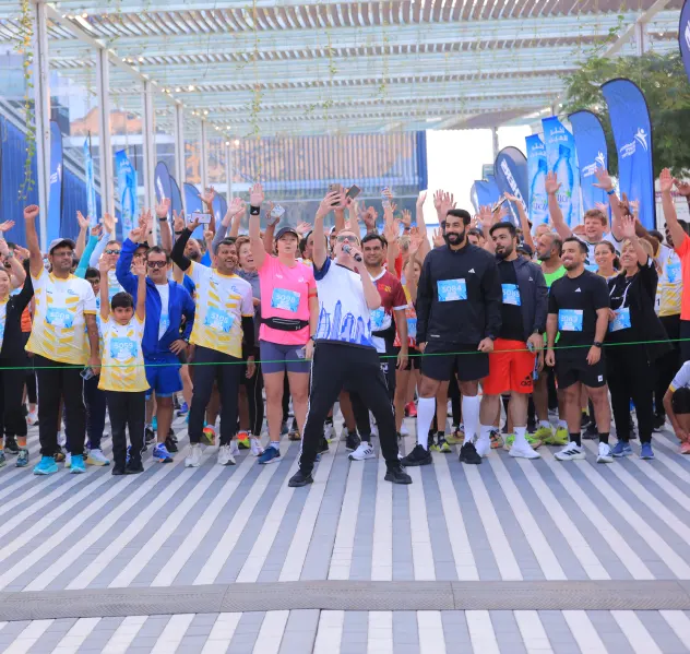 A diverse group of runners celebrating at a race start line, with many participants raising their hands in excitement under banners.