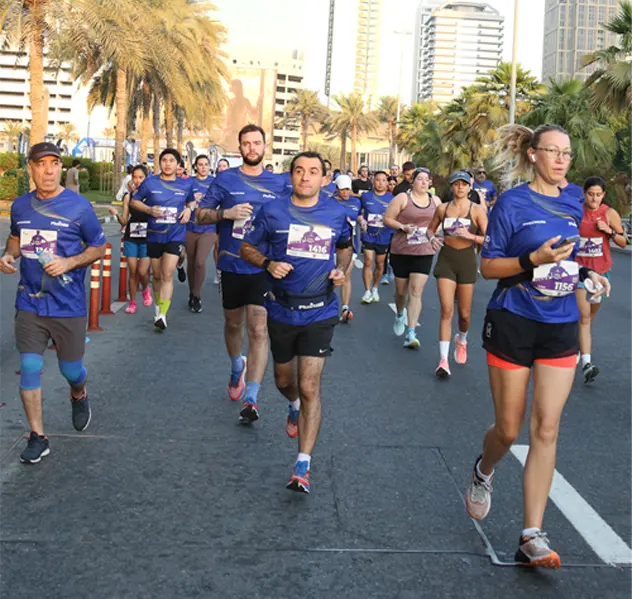 A group of runners in blue shirts races along a palm-lined street, showcasing a vibrant running event in an urban setting.