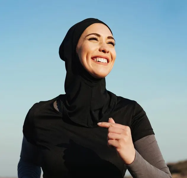 A person wearing a black athletic outfit and a head covering jogs outdoors against a clear blue sky.