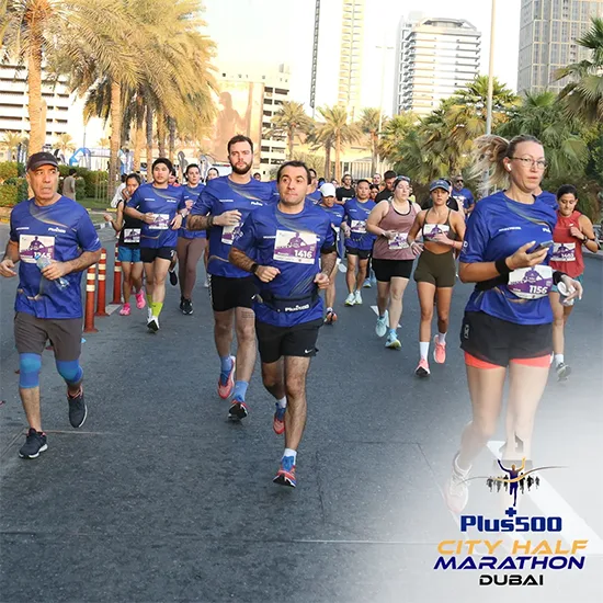 A diverse group of runners in blue shirts participate in the Plus500 City Half Marathon in Dubai, surrounded by palm trees and skyscrapers.