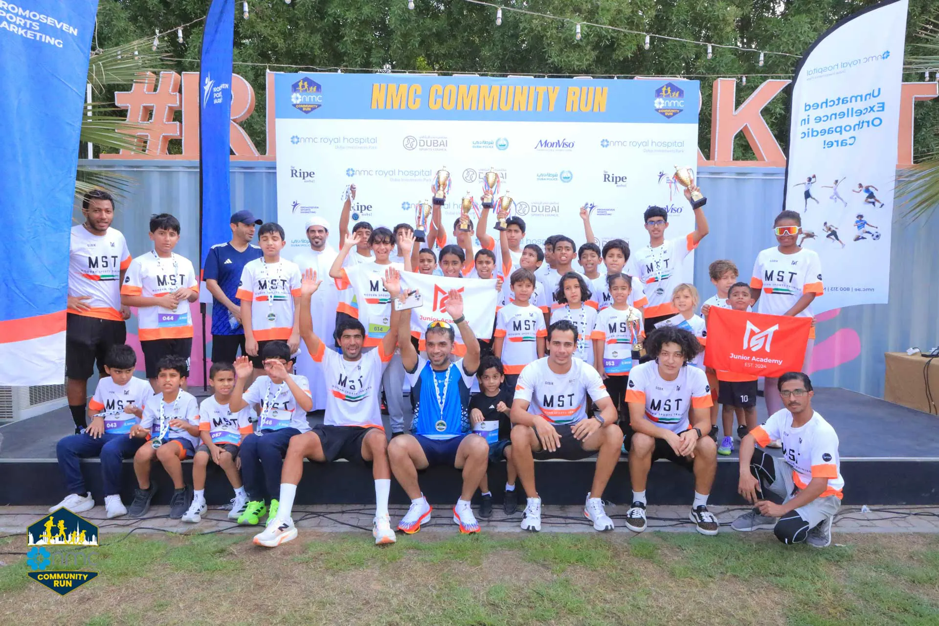 Group of participants celebrating at the NMC Community Run, displaying trophies and team banners on stage.