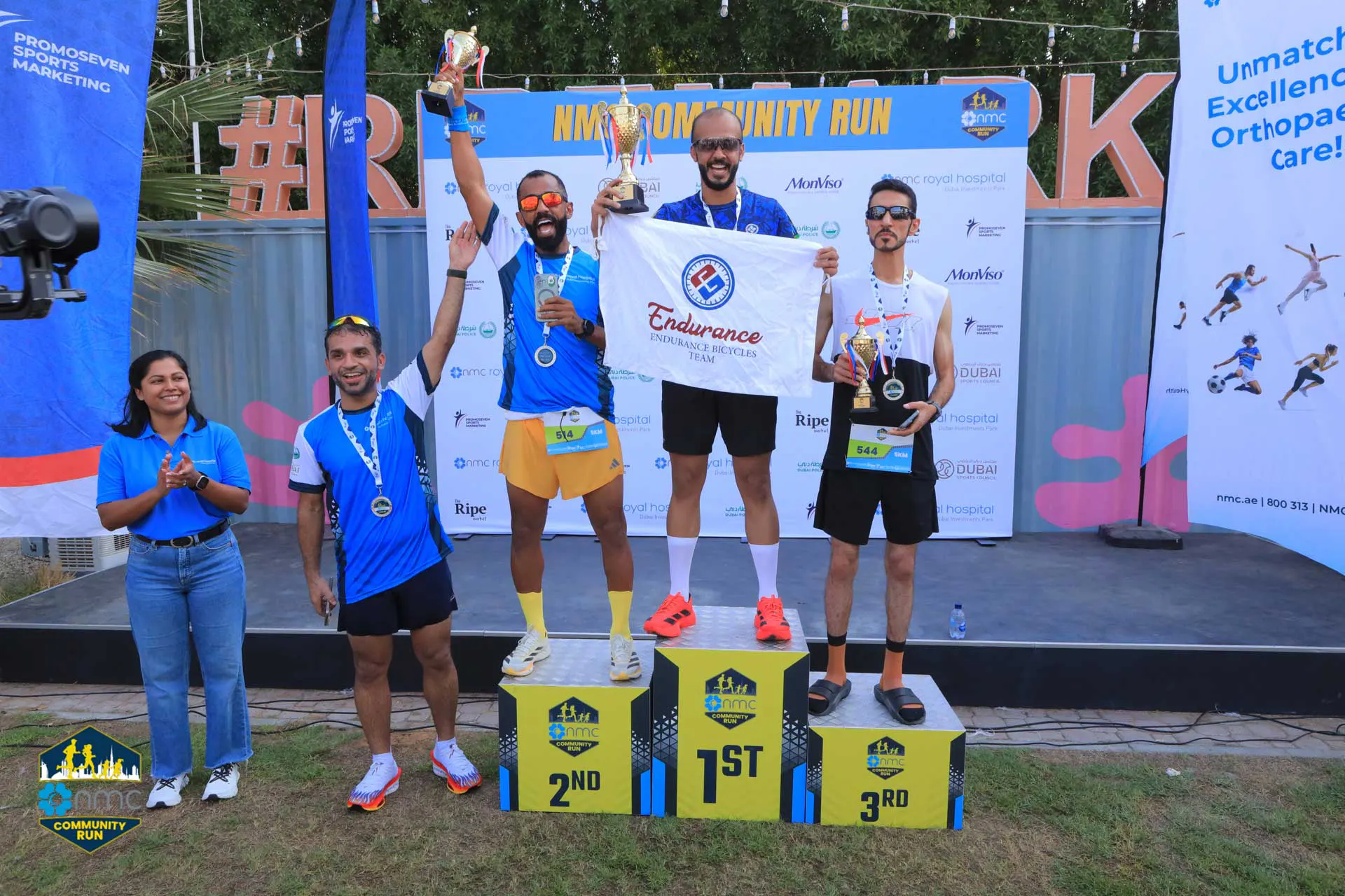 Participants celebrate on podiums at the NMC Community Run, holding trophies and a banner representing the Endurance team.