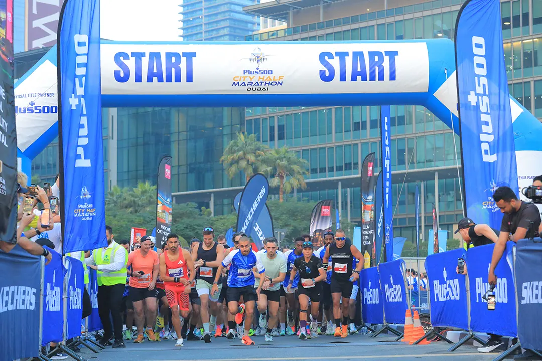 Runners sprint off the starting line at the Plus500 City Half Marathon in Dubai, with banners and spectators in the background.
