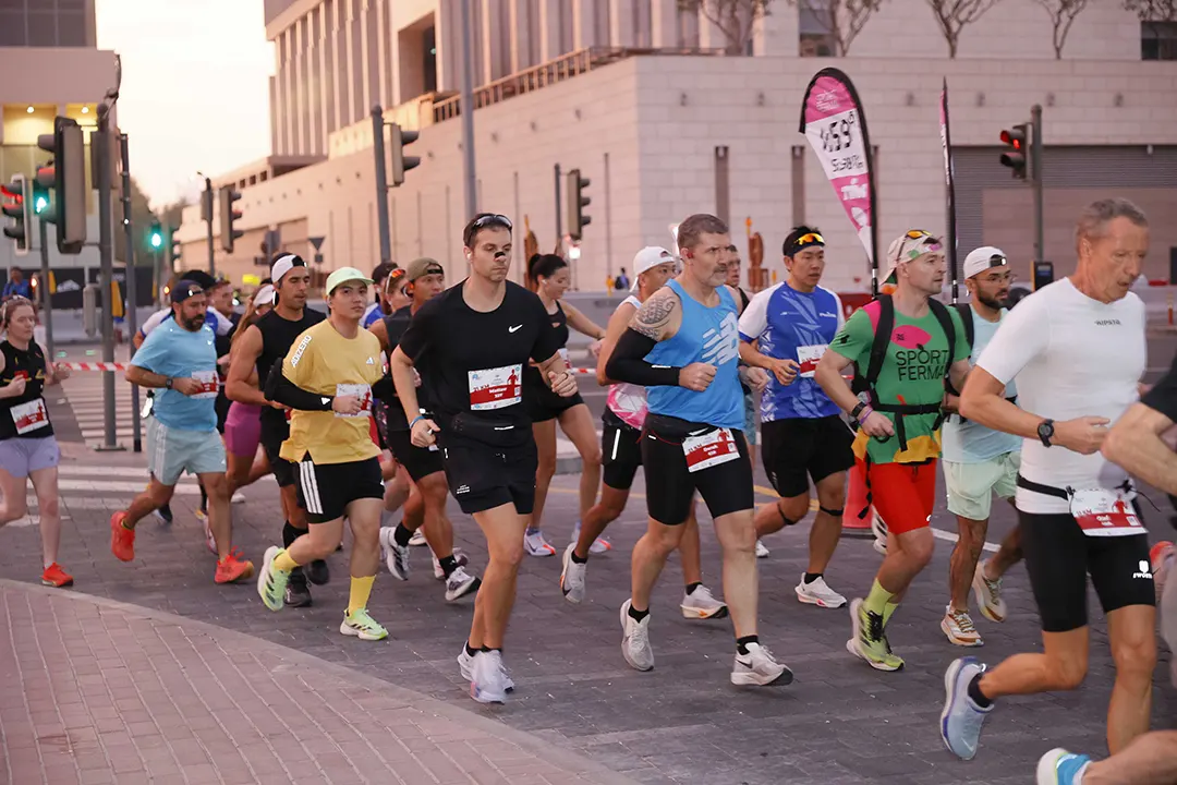 A diverse group of runners in athletic gear participate in a city race at dusk, with traffic lights and urban buildings in the background.