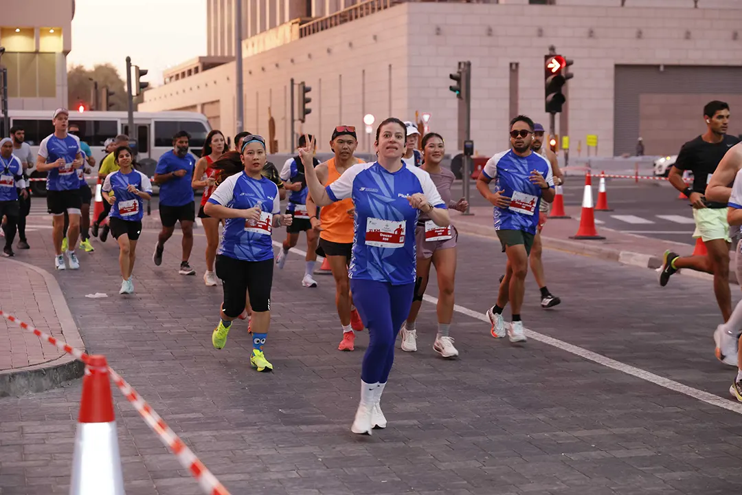 A group of runners in blue jerseys participates in a race on a city street, with traffic signals and road markers visible.
