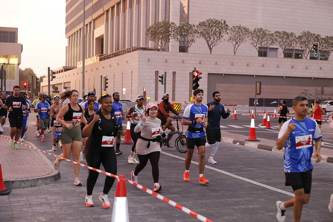 Runners participate in a city marathon, surrounded by traffic signals and cones, with buildings looming in the background at dusk.