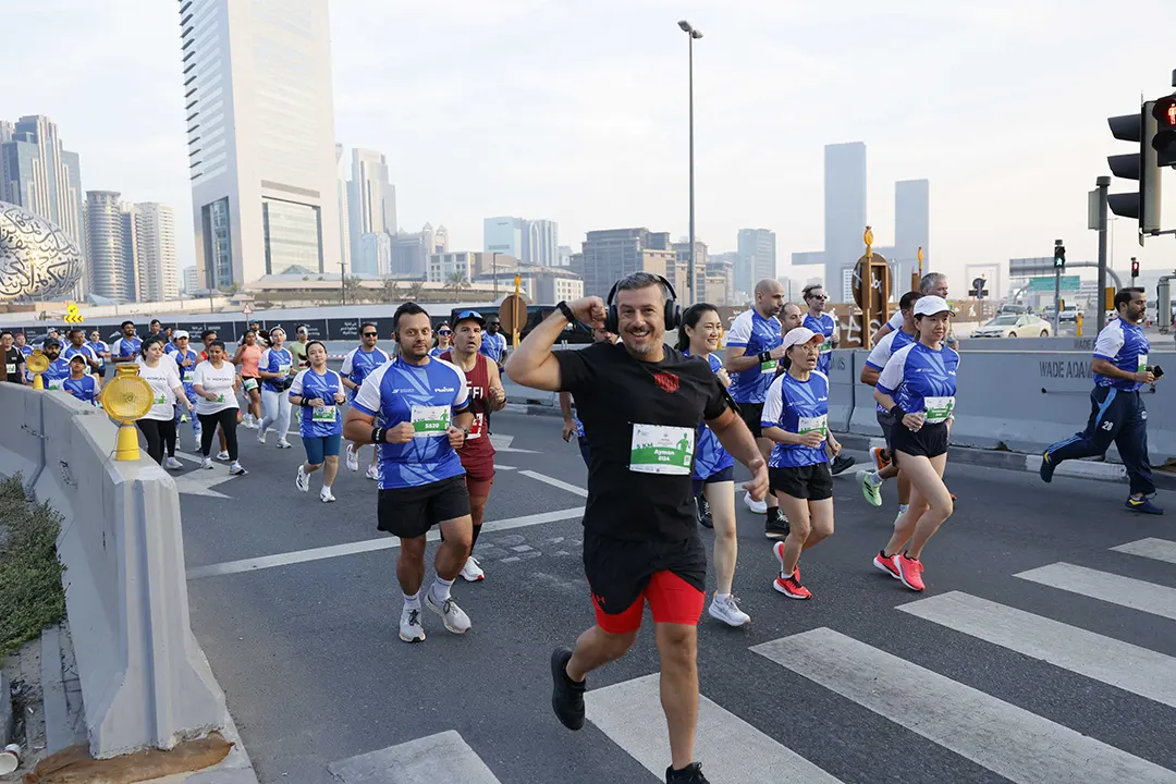 A vibrant group of runners in blue shirts participate in a city marathon, with urban skyscrapers visible in the background.