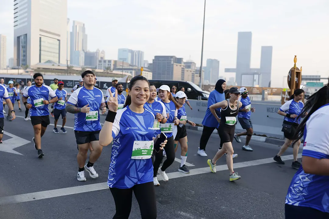 A group of runners in blue athletic shirts participate in a race on a city street, with buildings in the background.
