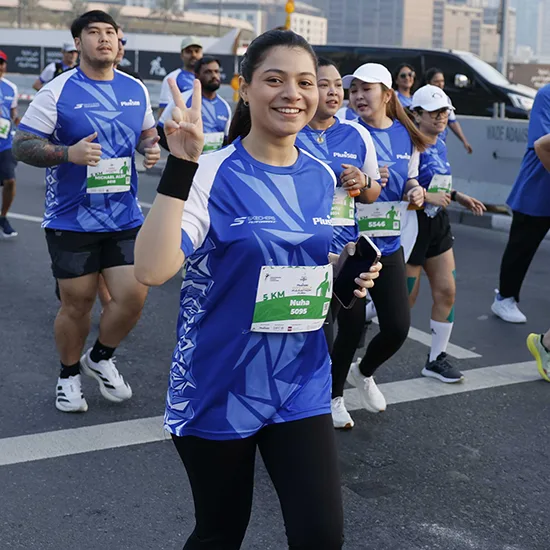 A group of runners wearing blue jerseys participates in a race, with one giving a peace sign while others jog alongside her.