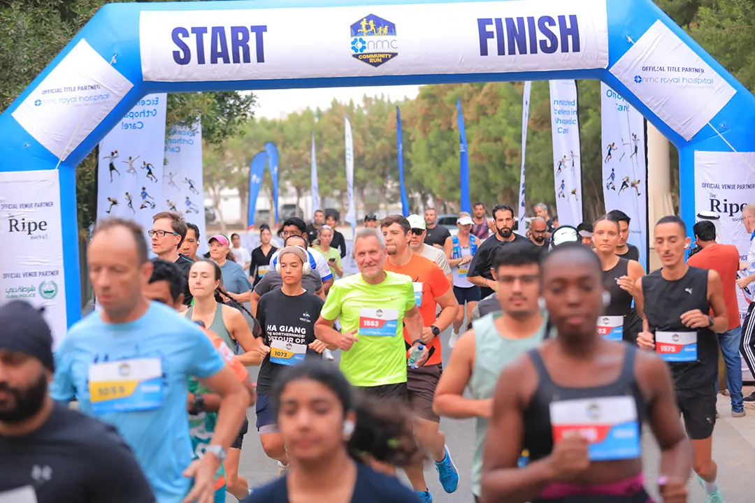 Runners enthusiastically participate in a community race, passing under a colorful start-finish archway amidst banners and trees.