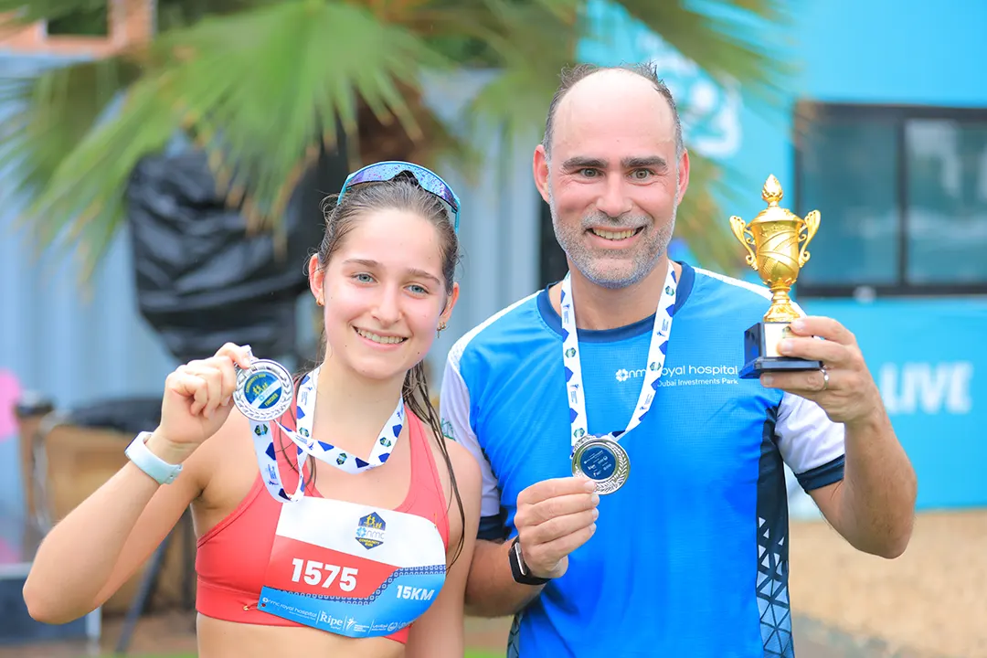 Two athletes proudly display their medals after a race, with one holding a trophy, set against a tropical backdrop.