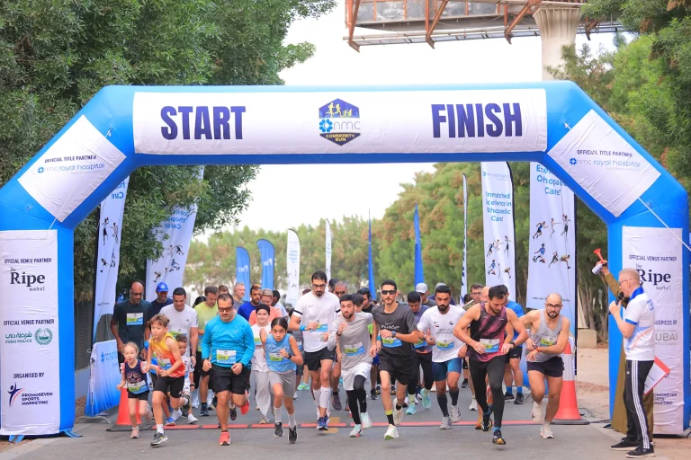 Participants sprint through an inflatable start and finish arch at a community run event, surrounded by banners and trees.