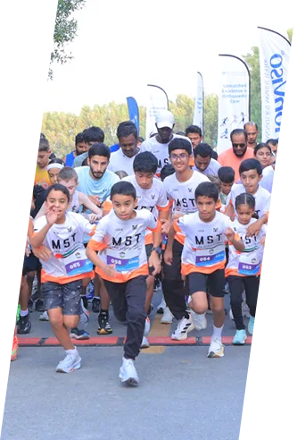 A group of children in matching shirts sprints energetically at the start of a race, with banners and spectators in the background.