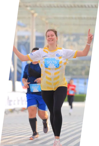 A runner wearing a yellow and white shirt celebrates with arms raised during a race, with another participant in blue behind.